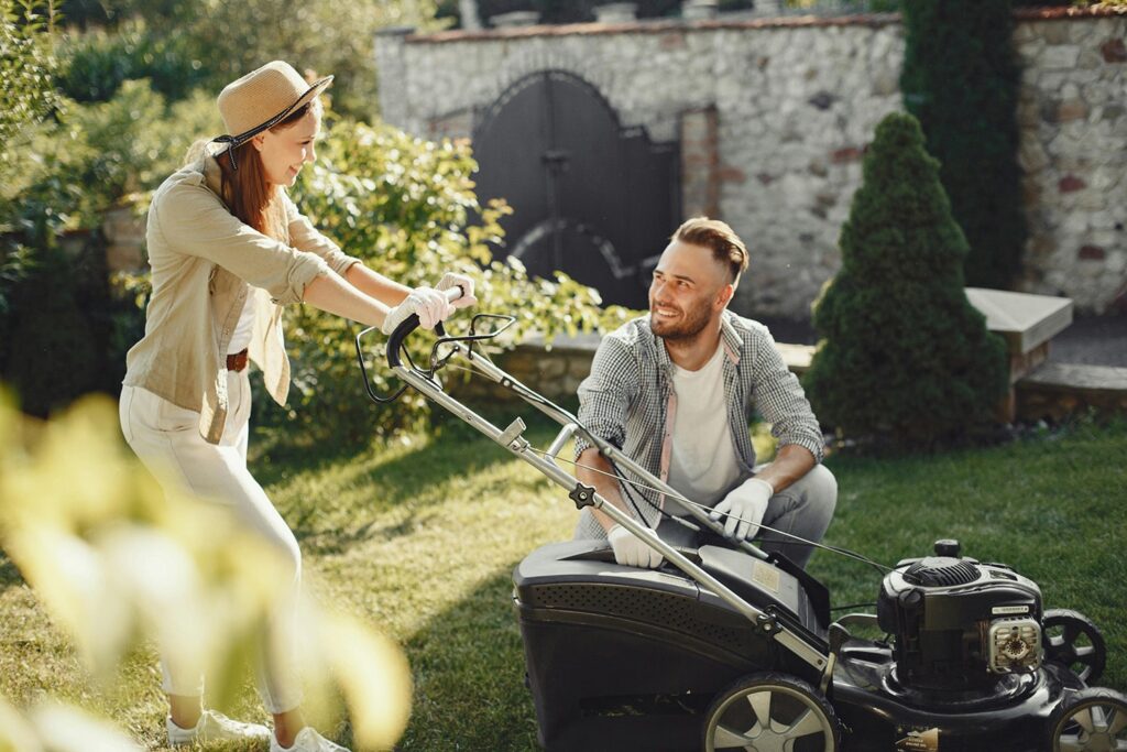 A man and woman are outside, the woman is pushing a lawn mower.