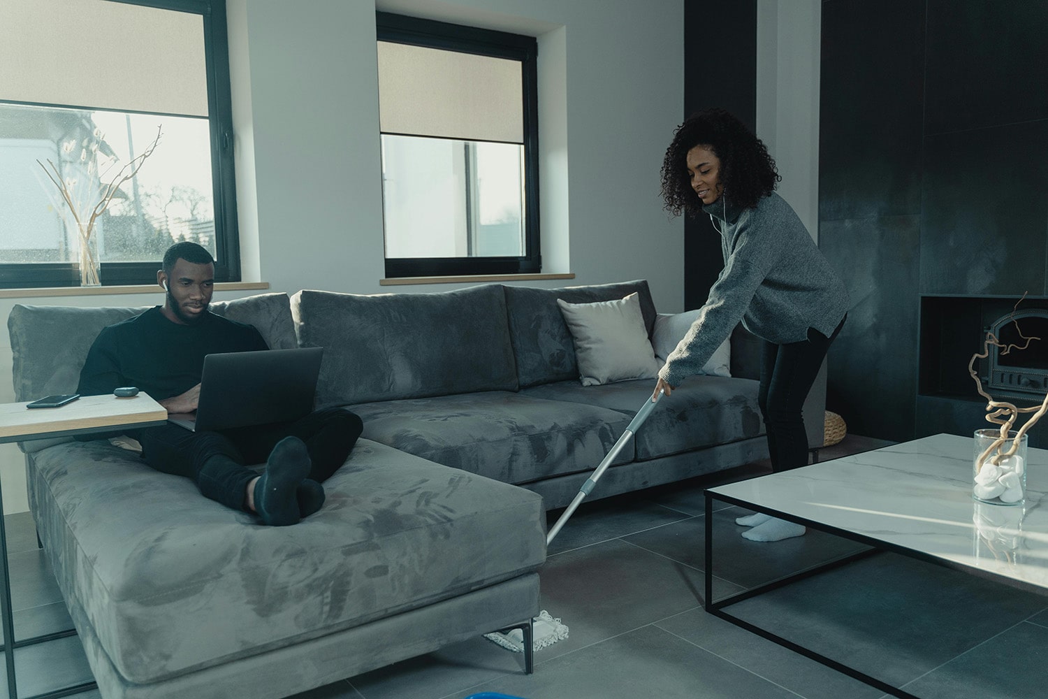 A woman is cleaning the floor, while a man sits on the sofa.